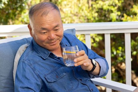 Senior asian man relaxing on porch chair sipping lemon water on sunny deck with greenery