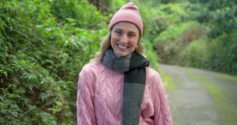 Smiling woman standing on woodland trail wearing pink cable knit sweater and gray scarf