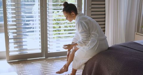 Woman Applying Lotion to Leg in Bright Bedroom with Shutters