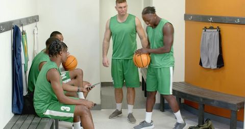 Locker room basketball team bonding with diverse players sharing phone and passing ball