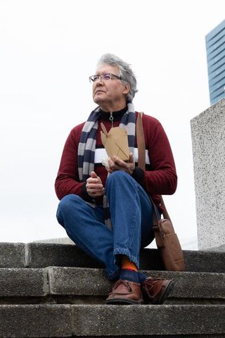 Senior man sitting on city steps eating takeout, wearing scarf and glasses