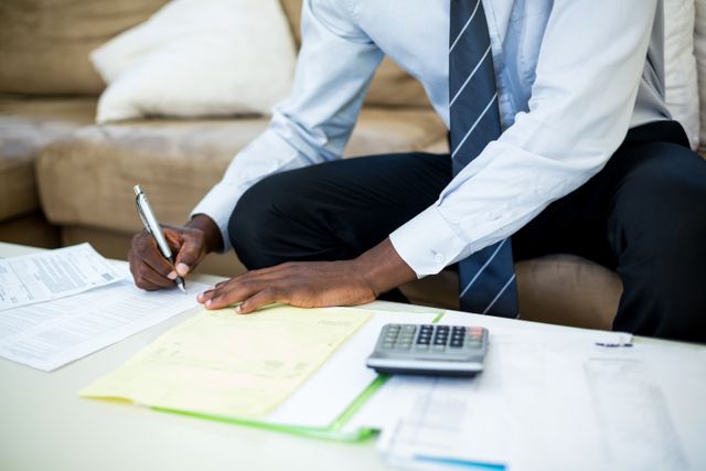 Man sitting on sofa managing finances, working on bills and paperwork with calculator and pen. Useful for themes related to home finance management, budgeting, personal accounting, and financial planning.