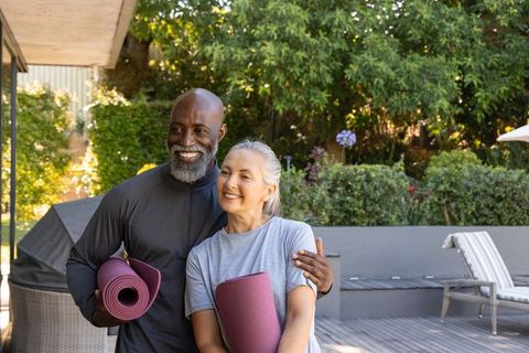 African american man and senior woman holding yoga mats on sunlit backyard deck smiling
