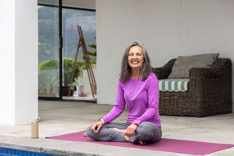 Senior woman practicing yoga on patio by pool, smiling in purple top and gray pants