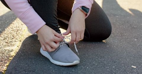 Woman kneeling tying grey sneaker laces on sunlit asphalt smartwatch ring casual fitness