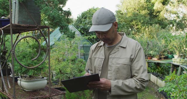 Senior man wearing cap and jacket writing on clipboard while standing in lush, green garden surrounded by plants and foliage. Useful for topics on gardening supplies, senior activities, working in nature, horticulture, plant care, greenhouse maintenance, and ecological preservation.