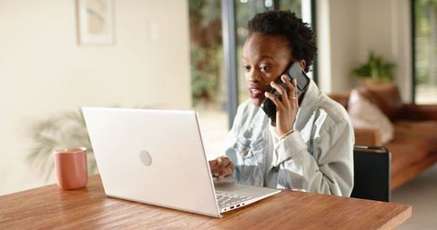 African american woman multitasking on phone and laptop in sunlit cozy home office