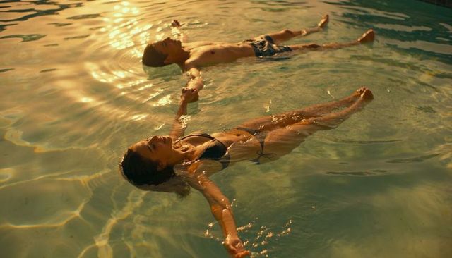 Couple floating, holding hands in sunlit pool at golden hour, romantic summer relaxation
