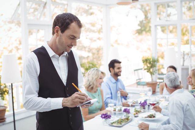 Smiling waiter writing order on notepad while friends dining in background