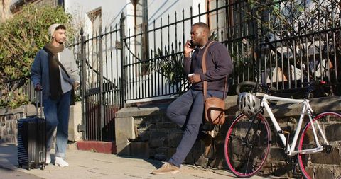 City commuters talking on phone while leaning on wall with bicycle, luggage, coffee