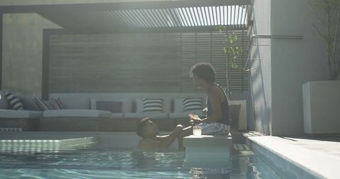 Sunlit poolside romance between two men holding hands at luxury patio pool by pergola