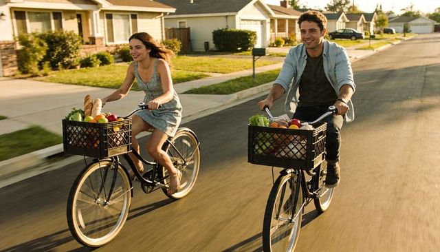 Young couple riding bicycles with grocery milk-crate baskets on sunlit suburban street