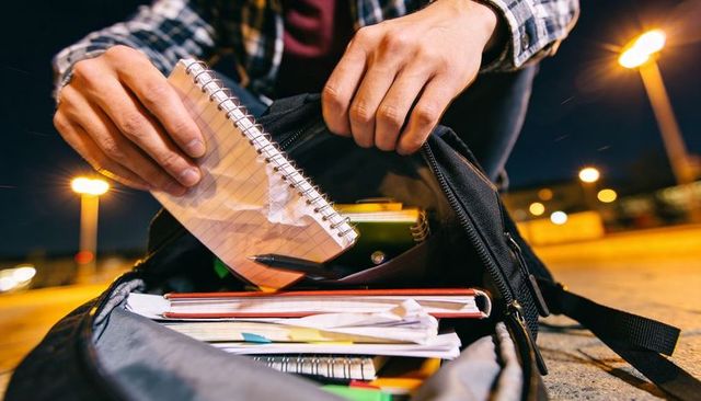 Student placing spiral notebook into backpack on urban pavement at night with bokeh