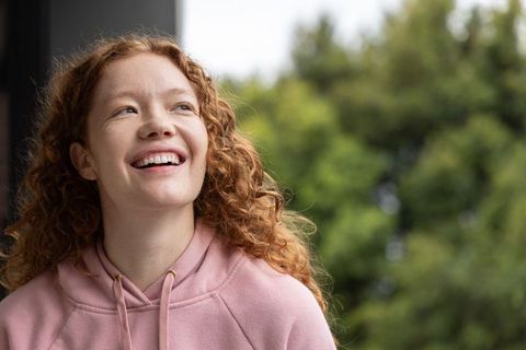 Smiling woman in her twenties wearing pink hoodie looking upward on porch with green bokeh