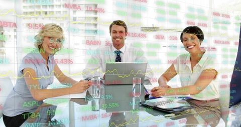Smiling business team collaborating around laptop with stock ticker overlays in glass boardroom