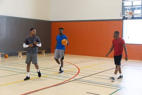 African american coach walking with two players holding basketball on indoor gym court