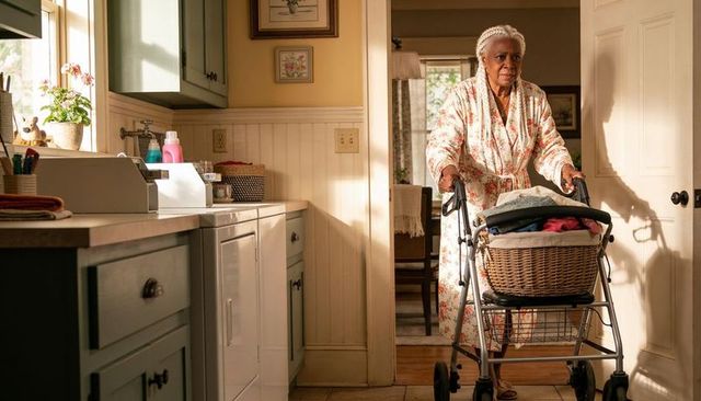 Senior woman pushing rollator with laundry basket in sunlit kitchen steadying independence