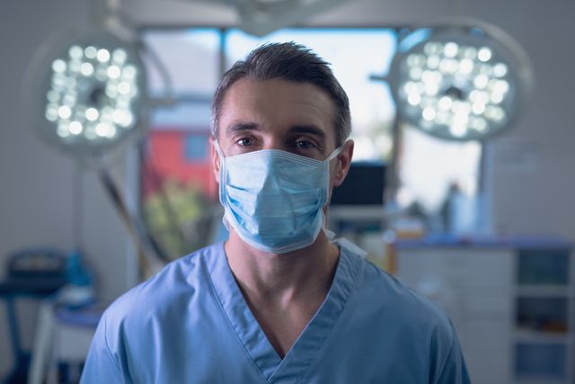 Male surgeon standing in an operating room wearing a surgical mask. Ideal for use in medical, healthcare, and hospital-related content. Useful for articles on healthcare professionals, medical practices, surgical procedures, and hospital environments.