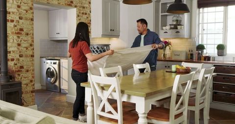 Couple spreading tablecloth in cozy farmhouse kitchen showing teamwork and rustic dining