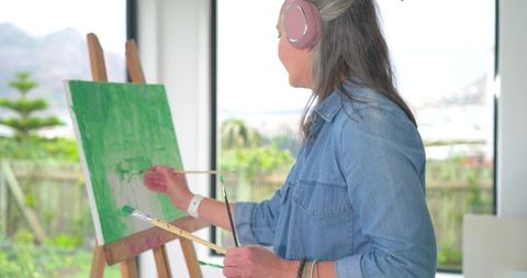 Mature woman painting green canvas wearing denim shirt and pink headphones