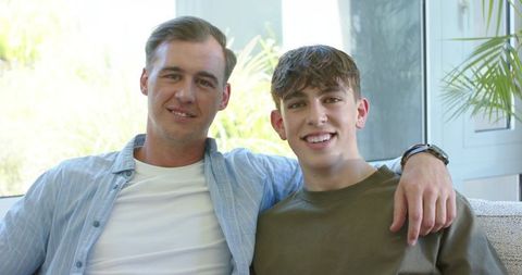 Father and teen son relaxing on sofa smiling in sunlit living room with greenery