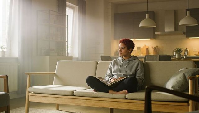 Young woman reading on sofa in sunlit minimalist scandinavian living room, calm lifestyle