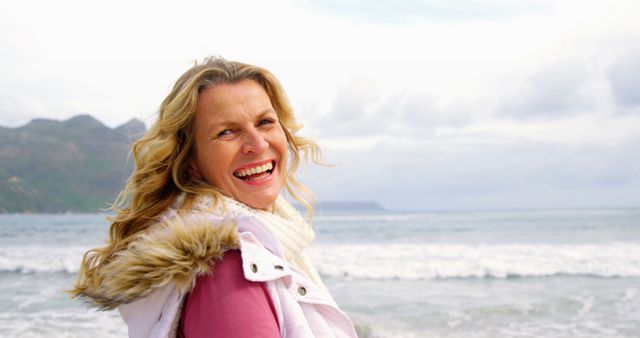 Woman with long blonde hair smiling and enjoying the beach on a cloudy day. Ocean waves and distant mountains visible. Suitable for use in themes related to happiness, outdoor activities, mental wellness, nature, vacations, and lifestyle.