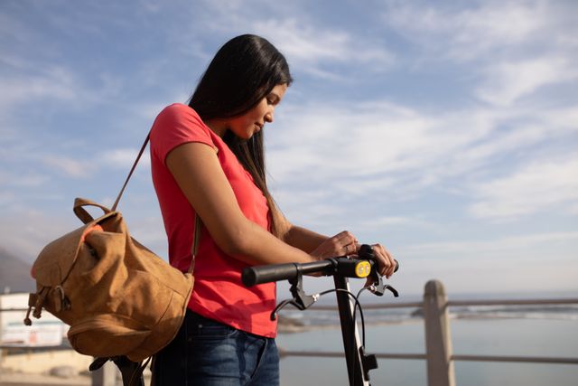 Side view of a biracial woman enjoying free time in nature on a sunny day, riding a scooter, wearing beige backpack, adjusting at the smartwatch.