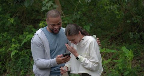 Laughing black man and white woman checking smartphone on forest trail, outdoor moment