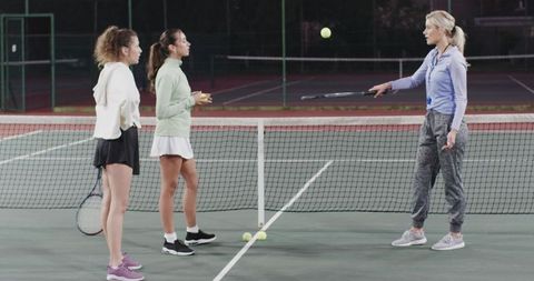 Female tennis coach training teen players on floodlit outdoor court