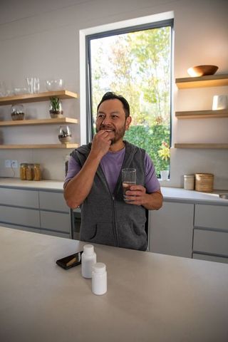 Mature asian man taking daily vitamins at modern kitchen island holding glass of water
