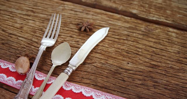 Old-fashioned cutlery set with fork, spoon, and knife placed on a rustic wooden table. Perfect for illustrating kitchen decor blogs, antique themed dining settings, or farmhouse lifestyle articles. Red lace trim adds a touch of elegance and warmth to the homely setup. Suitable for use in food and dining advertisements, culinary presentations, and hospitality industry promotions.