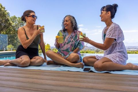 Three women relaxing by infinity pool sharing citrus drinks on sunny terrace, friends laughing