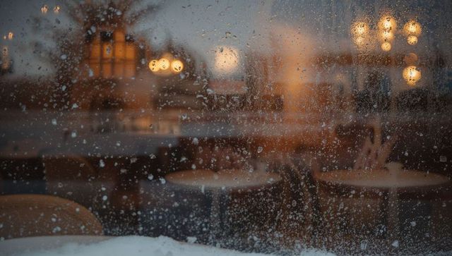 Rain-dotted cafe window framing warm bokeh lights and snow-covered sill at dusk