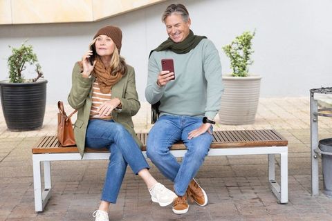 Mature couple sitting on bench using smartphones in urban plaza, casual outdoor lifestyle
