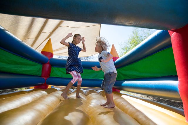 Two happy siblings are jumping on a bouncy castle at a playground. They are enjoying their time together, showcasing the joy and energy of childhood. This image can be used for promoting family activities, playgrounds, summer camps, and children's events. It highlights the fun and carefree nature of outdoor play.