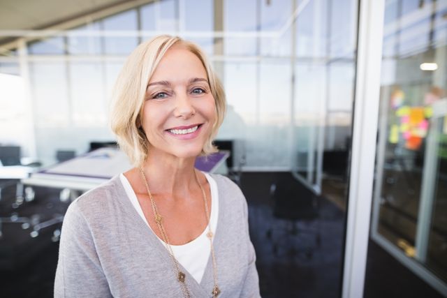 Mature businesswoman smiling confidently while standing in a modern office environment. Ideal for use in corporate websites, business presentations, career-related articles, and professional networking profiles.