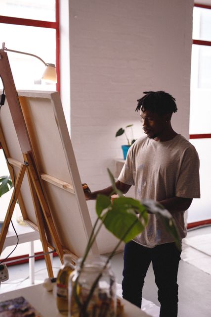This stock photo depicts an African American male painter concentrating on his work in an art studio. He is painting on a large canvas, surrounded by art supplies. This image can be used for articles and designs related to creativity, artistic expression, hobbies, and the work environment of artists. It showcases the dedication and focus involved in the creative process.
