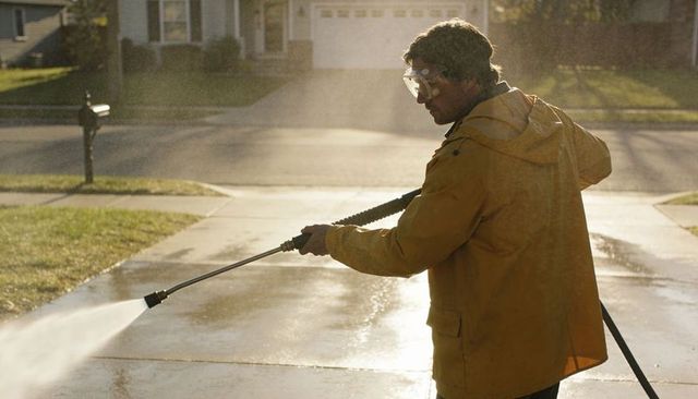 Backlit man pressure washing suburban driveway wearing yellow jacket and safety goggles