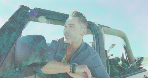 Mature man leaning on open-top jeep during sunlit coastal road trip with guitar