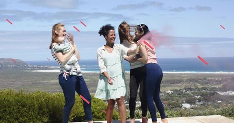 Laughing friends hugging and holding baby on sunlit coastal terrace overlooking ocean