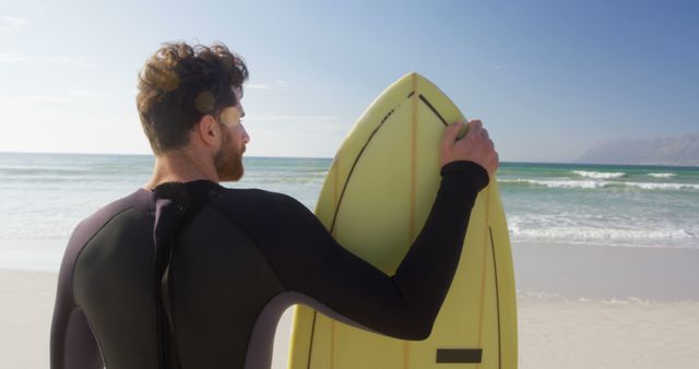 A young Caucasian man in a wetsuit holds a surfboard on the beach, gazing towards the ocean, with copy space. His anticipation for the surf session ahead is palpable against the backdrop of the serene coastline.