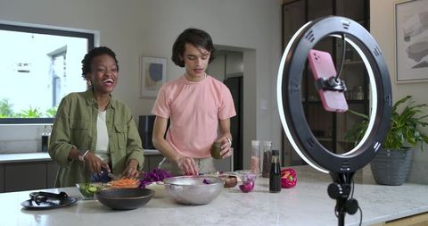 Two friends filming cooking tutorial while chopping vegetables at kitchen island