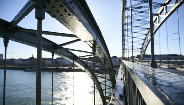 Wet steel truss bridge with arched spans and rivets over sunlit river reflecting city skyline