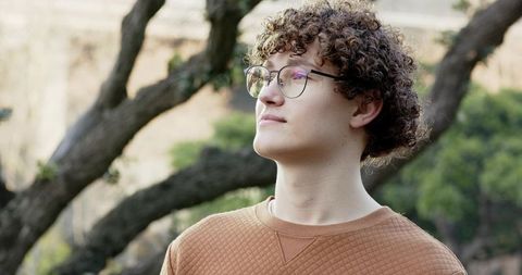 Young man gazing upward in park wearing round glasses and rust-brown quilted sweatshirt