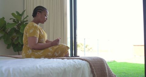 African american woman sitting on bed holding phone in sunlit minimalist bedroom