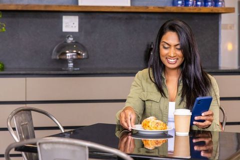 Indian woman enjoying coffee and croissant while using smartphone at glossy cafe table, smiling
