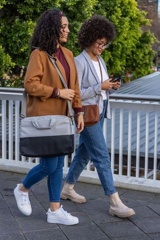 Two friends walking urban bridge in blazers and jeans with tote and crossbody bags