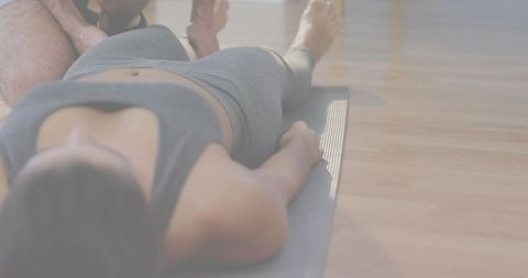Woman lying on yoga mat doing core exercise with trainer assisting on wooden floor, desaturated ligh