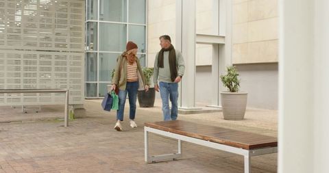 Mature couple walking in urban courtyard carrying reusable bags and wearing scarves
